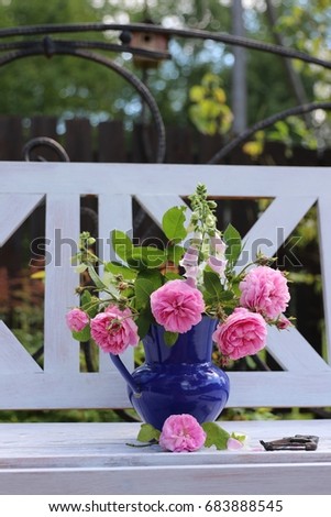 Roses, foxglove in vintage dark blue enamelware jug, rustic secateurs on white bench, summer morning in garden, floral composition, daylight, vertical photo