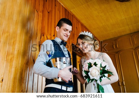 A stylish happy groom with a butterfly keeps a bucket, pail of ice and champagne. Bride with a bouquet of flowers Peonies. On the background of a wooden wall and a picture. Wedding Morning. Close up