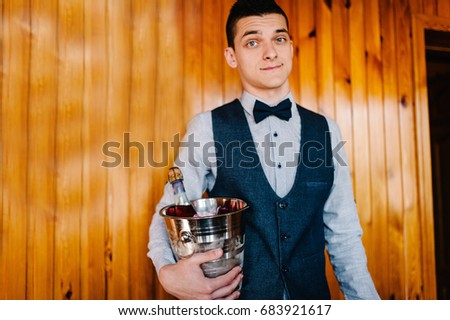 A stylish happy groom with a butterfly keeps a bucket, pail of ice and champagne. On the background of a wooden wall and a picture. Wedding Morning. Close up