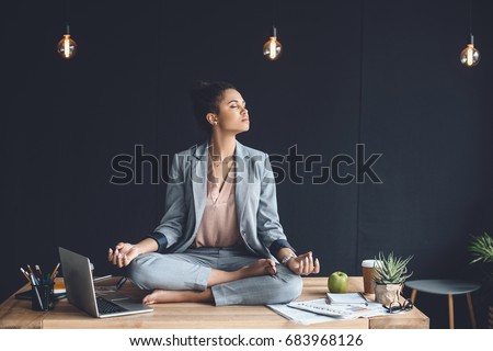 african american businesswoman sitting in lotus pose on table while meditating in office Royalty-Free Stock Photo #683968126