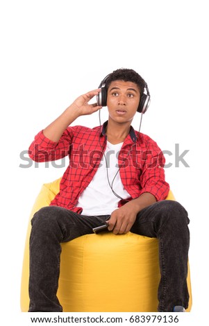 Happy friendly teenage boy with headphones sitting on a yellow chair and listening to music, teenager wearing red shirt,  isolated on white background