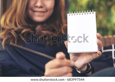Closeup image of a beautiful Asian woman holding and showing blank notebook in office with green vertical garden background