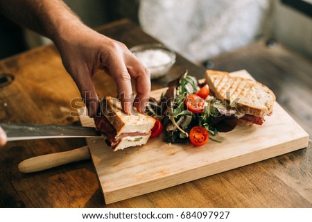 Man prepares lunch, serves grilled bread sandwich snack on top of wooden cutting board, with side of green salad, healthy alternative to burgers and grease Royalty-Free Stock Photo #684097927