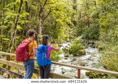 New zealand travel tourists taking phone picture on tramping hike in forest with backpacks. Woman holding smartphone taking photos of river on Routeburn track hiking trail on South Island.