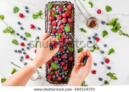 Chocolate cake tart with chocolate cream, woman decorate it with blueberry raspberry mint leaves, powdered sugar. copy space top view hands in picture
