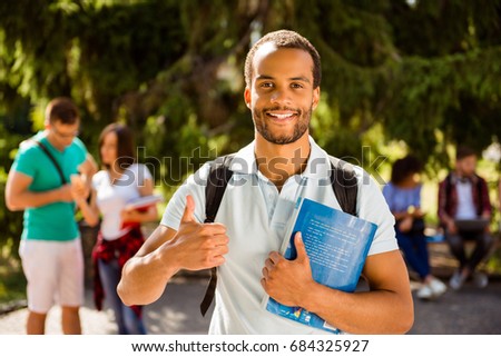 Young cheerful successful african nerdy student is showing thumb up, standing with books, his classmates on the background, park near campus, sunny day