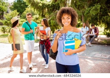 Portrait of an attractive young student, standing with rucsack and note pads, with beaming smile, behind are her friends, talking outside in a sunny park near college