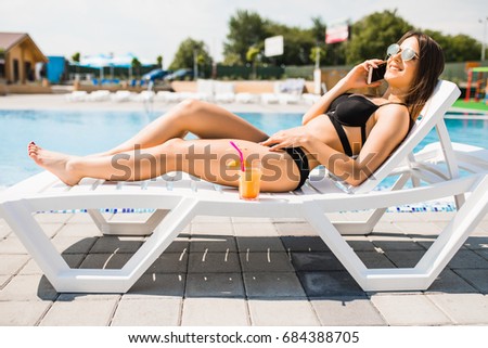 Young woman in swimsuit relaxing with cocktail on chaise lounge and talk on phone . Time summer photo.