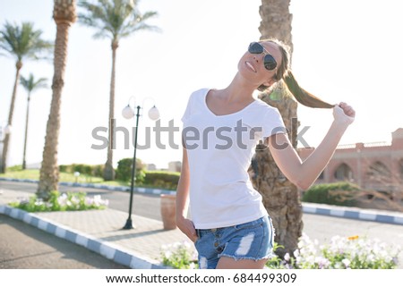 Photo of style pretty woman in the sunlight on the background road with palm trees. She has fun. She dressed in white t-shirt and black sunglasses. Vacation mood. Sunny summer day.