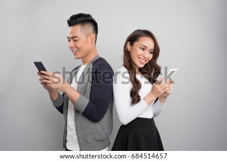 Beautiful young asian couple holding mobile phones and standing back to back against grey background Royalty-Free Stock Photo #684514657
