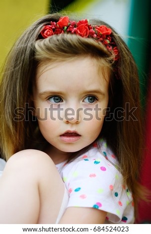 Portrait of the charming girl of 4-5 years. Beautiful long fair hair. A wreath from red colors on the head. Expressive gray-blue eyes. Bright lips.