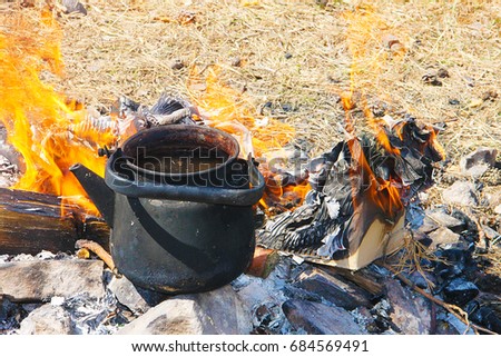 A black smoked teapot stands on a fire surrounded by yellow flame tongues against the background of dry grass - tourist inventory in the trek.