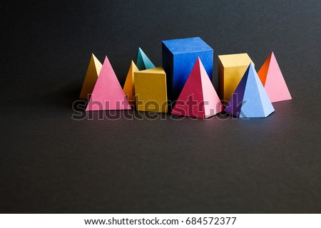 Colorful abstract geometric solid figures on black background. Pyramid prism rectangular cube yellow blue pink green colored figures. Black textured paper, shallow depth of field, copy space.