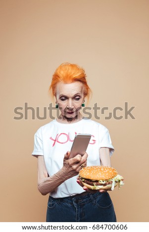 Picture of attractive mature redhead fashion woman standing isolated at studio make photo of burger by phone. Looking aside.