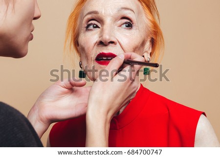 Picture of gorgeous mature redhead fashion woman standing isolated at studio near makeup artist doing makeup of her lips. Looking aside.