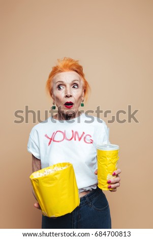 Picture of shocked mature redhead fashion woman standing isolated at studio eating popcorn and drinking aerated water. Looking aside.