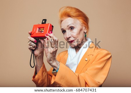 Picture of mature redhead fashion woman standing isolated at studio and holding camera. Looking aside.