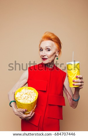 Picture of smiling mature redhead fashion woman standing isolated at studio eating popcorn and drinking aerated water. Looking camera.