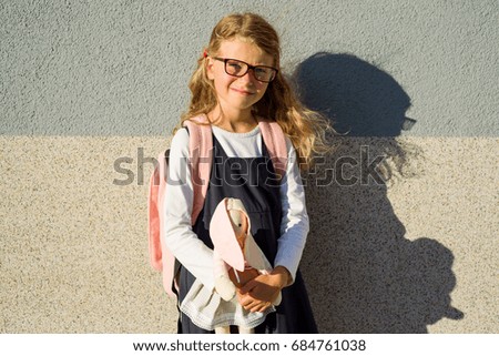 A cute schoolgirl of primary school is smiling, holding  a toy. Girl with a backpack near the building outdoors. Leaving childhood and  Back to school!
