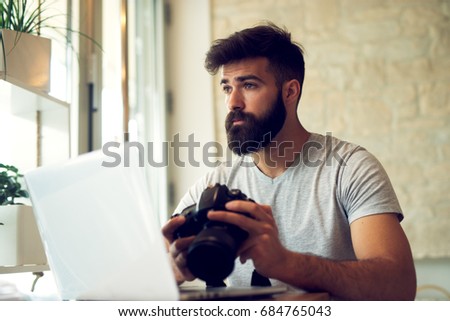 Portrait of young photographer holding camera sitting in front of laptop.