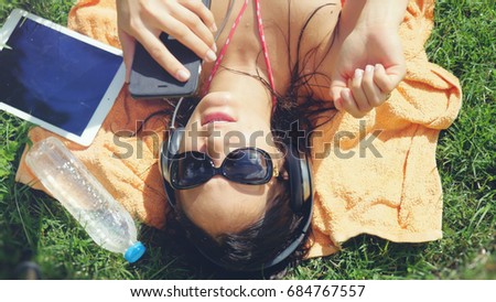 Beautiful young woman in sunglasses lying on the grass, listening to music and uses mobile phone