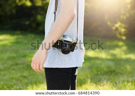 Cropped shot of male`s body with retro camera on shoulder standing at green grass going to photographing someone. Professional talented male photographer posing with vintage camera at greenland