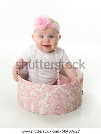 Portrait of an adorable baby girl sitting in a pink and white hatbox wearing a white shirt, pearl necklace, and pink headband with rose