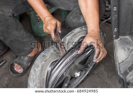 Man is changing old leaked tyre by taking off from alloy wheel with hands and tool in the dark garage where he's working.