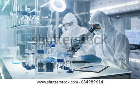 In a Secure High Level Laboratory Scientists in a Coverall Conducting a Research. Chemist Adjusts Samples in a  Petri Dish with Pincers and then Examines Them Under Microscope. Royalty-Free Stock Photo #684989614