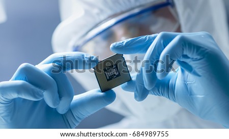 In Ultra Modern Electronic Manufacturing Factory Design Engineer in Sterile Coverall Holds Microchip with Gloves and Examines it. Royalty-Free Stock Photo #684989755