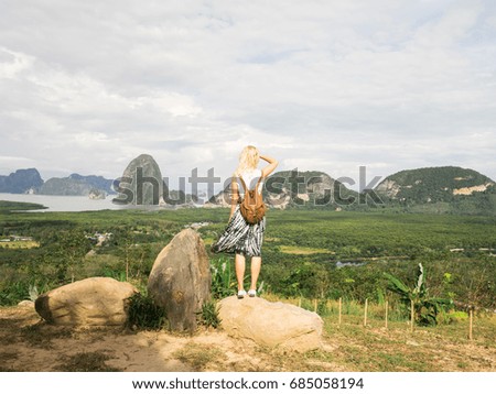Back view of girl with small backpack posing on rock looking at green valley and ocean. 