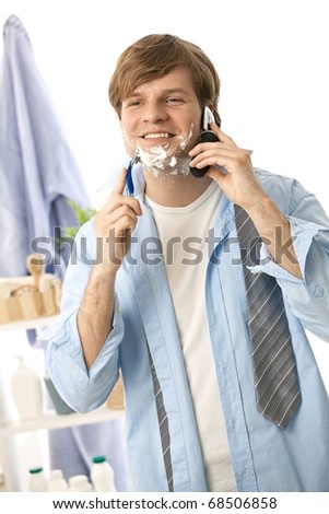 Young man talking on mobile while shaving. Isolated on white.