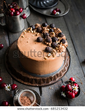 chocolate cake with cherries and nuts is decorated with mini-cakes, chocolate cheese cream and nuts on a wooden dark background. Fresh cherry and chamomile flowers