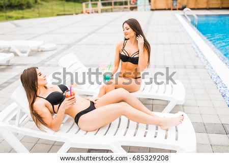 Two beautiful women are lying on chaise-lounge poolside outdoors and talking.