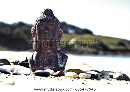 Selective focus meditating Buddha statue on sand in outdoor setting. Natural setting statue of Buddha at beach with pebble stones and burnt out, white sea waves, white sky, green hill in background.		