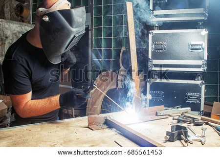 A bald, strong man in black work clothes   welds a metal welding machine  in a warehouse, against a background of a green glass wall, suitcases for equipment