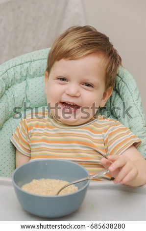 little blond boy in a striped T-shirt eats porridge with a spoon from a gray plate and sits in a nursery chair for feeding. He is cheerful, happy and well fed, laughing and fooling around