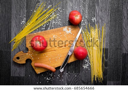 Top view of still-life picture with cutting board, fresh tomatos,sausage, spaghetti and knife surrounded by scattered rice on dark wooden background. Flat lay.