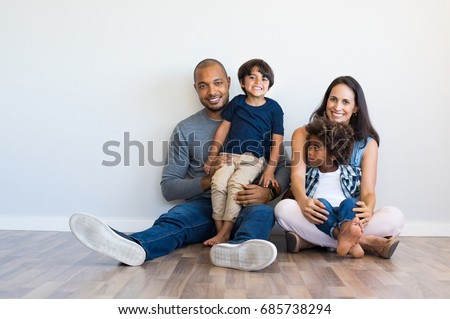 Happy multiethnic family sitting on floor with children. Smiling couple sitting with two sons and looking at camera. Mother and black father with their children leaning on wall with copy space. Royalty-Free Stock Photo #685738294