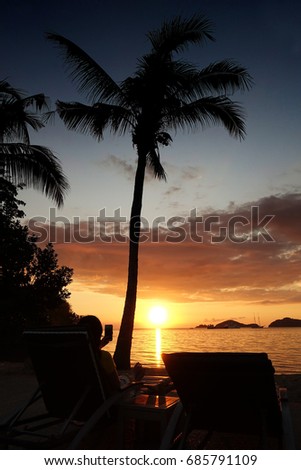 Silhouette young woman at beach laying on sunbed next to a palm tree enjoying watching and making picture with mobile phone of orange sun setting at night into the tropical ocean in Flores, Indonesia.