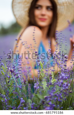Close-up of lavender flowers with a beautiful young woman on background 
wearing a straw hat standing in a big field of lavender plants. Vertical picture.