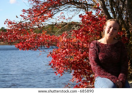 young woman is smiling. There are pond and tree with red leafs on background