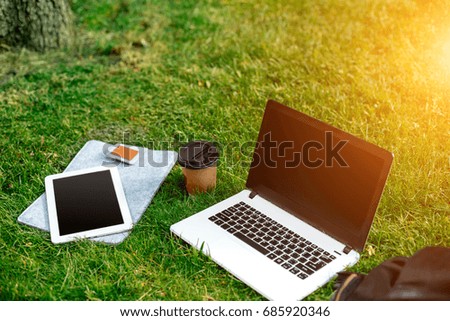 Laptop computer on green grass with coffee cup, bag and tablet in outdoor park