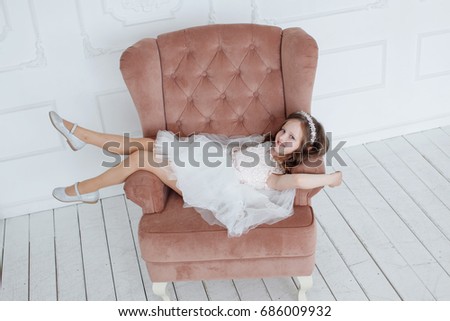 Beautiful little girl with curls and white Quinceanera dresses with a train lying in a pink seat and fun dangles her legs