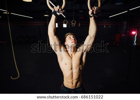 Young fit man pulling up on gymnastic rings. Bodybuilder in the gym is training, the dark background. Motivation. Looks up.