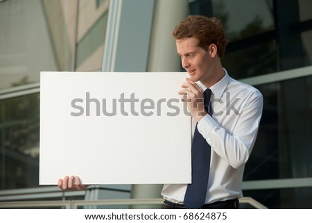 A cheerful businessman looking the side at blank poster sign outside of a glass office building exterior. Custom copy space. 20s handsome caucasian British male looking away