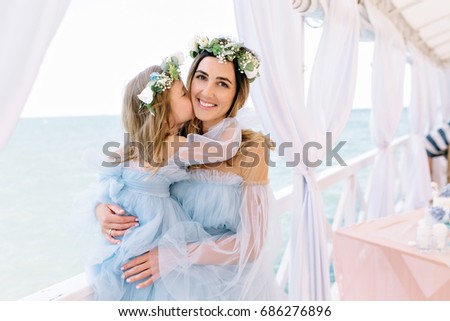 Picture of mother with little cute daughter wearing wreaths of flowers and blue dresses on summer tent. Girl kissed mother, mother is looking at camera with great smile. Birthday party.