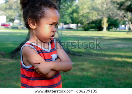 A young African American boy is unhappy at the park. His arms are crossed and his expressions shows he is angry or sad about something he sees.  Bullying concept, angry, sulking child Royalty-Free Stock Photo #686299105