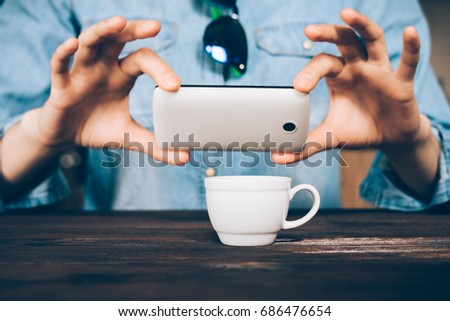 Woman in a denim shirt takes a picture of a cup of coffee at a cafe, close-up