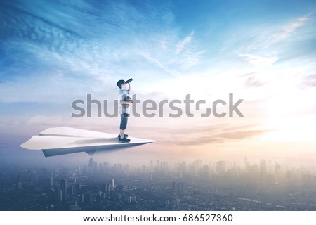 Picture of little boy standing on a paper plane while flying above city and looking with binoculars